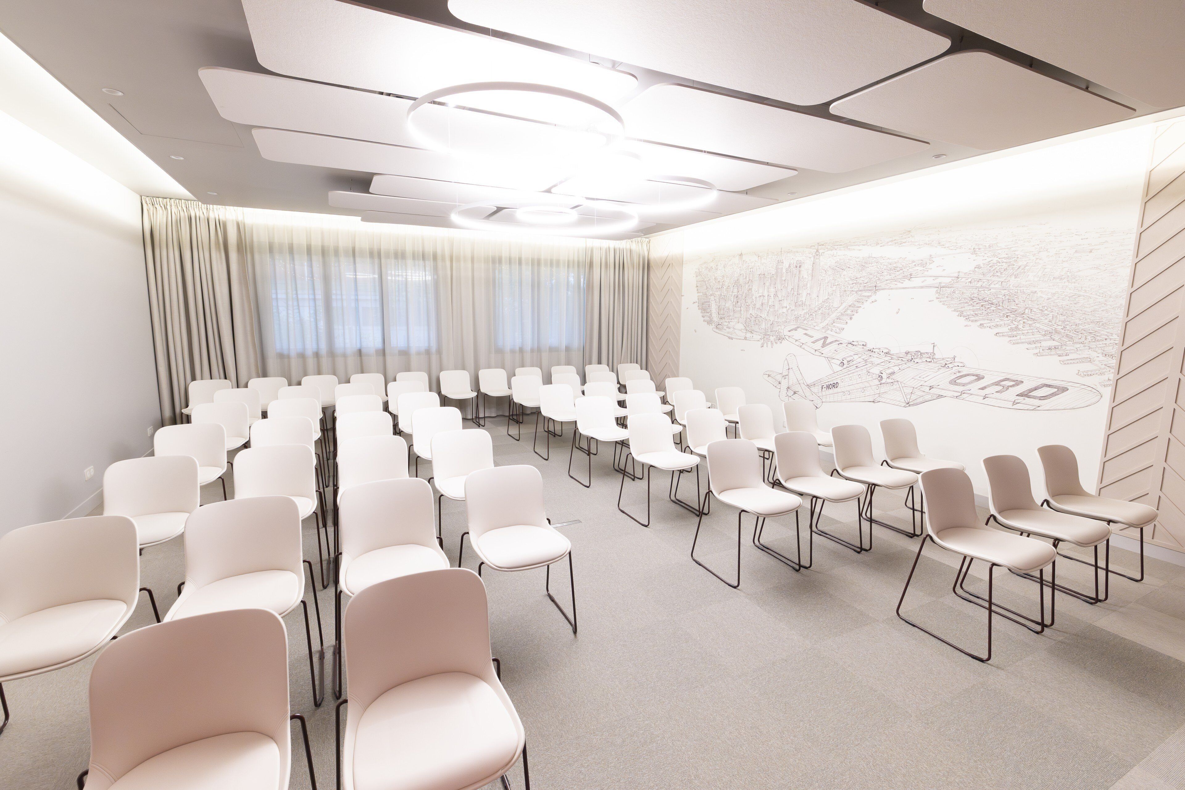 Una sala de conferències amb files de cadires blanques, un gran finestral amb cortines i un mural a la paret.