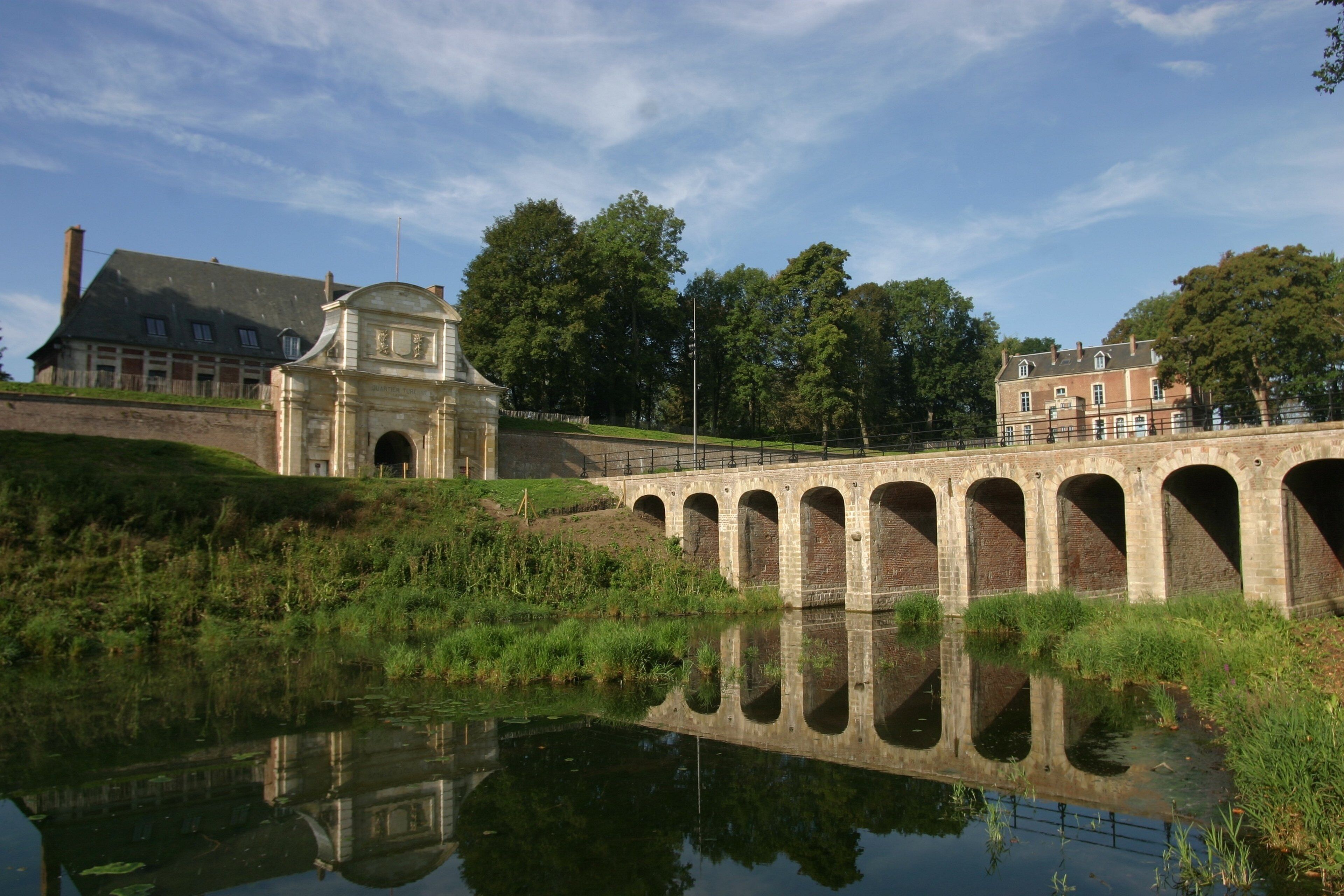 A stone bridge with multiple arches over a calm body of water, reflecting the surrounding trees and buildings.