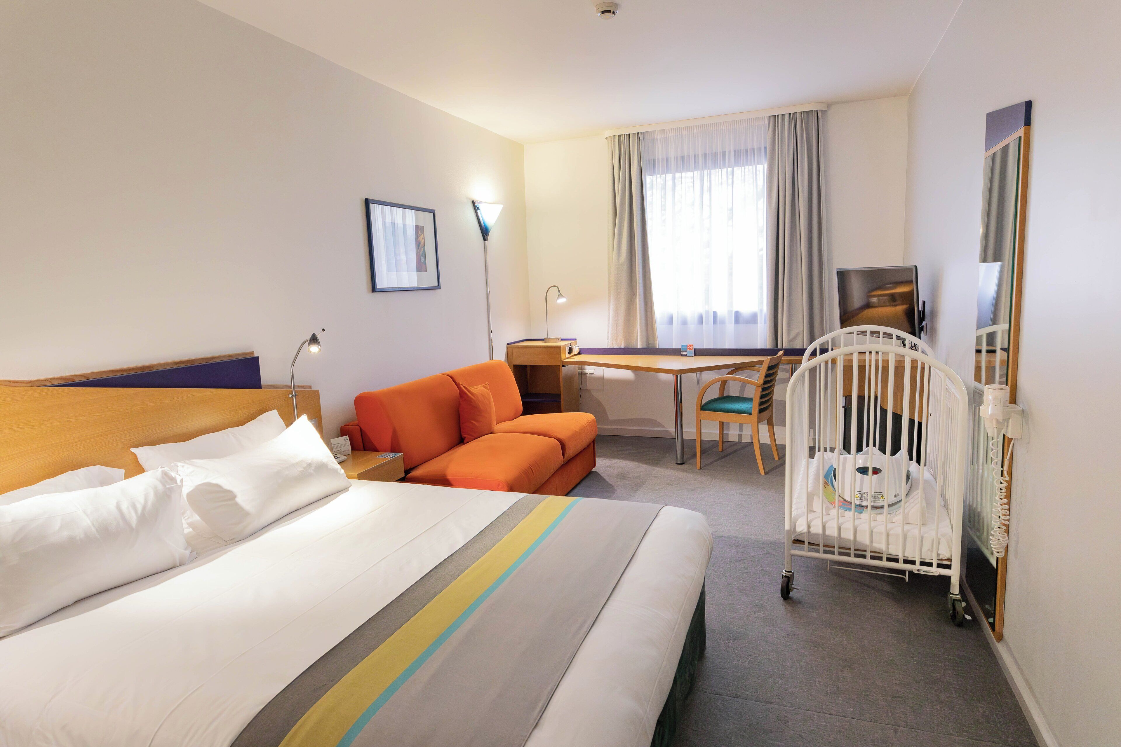 Une chambre d’hôtel comprenant un lit, un bureau, une chaise, une petite table, un berceau et une fenêtre avec des rideaux.