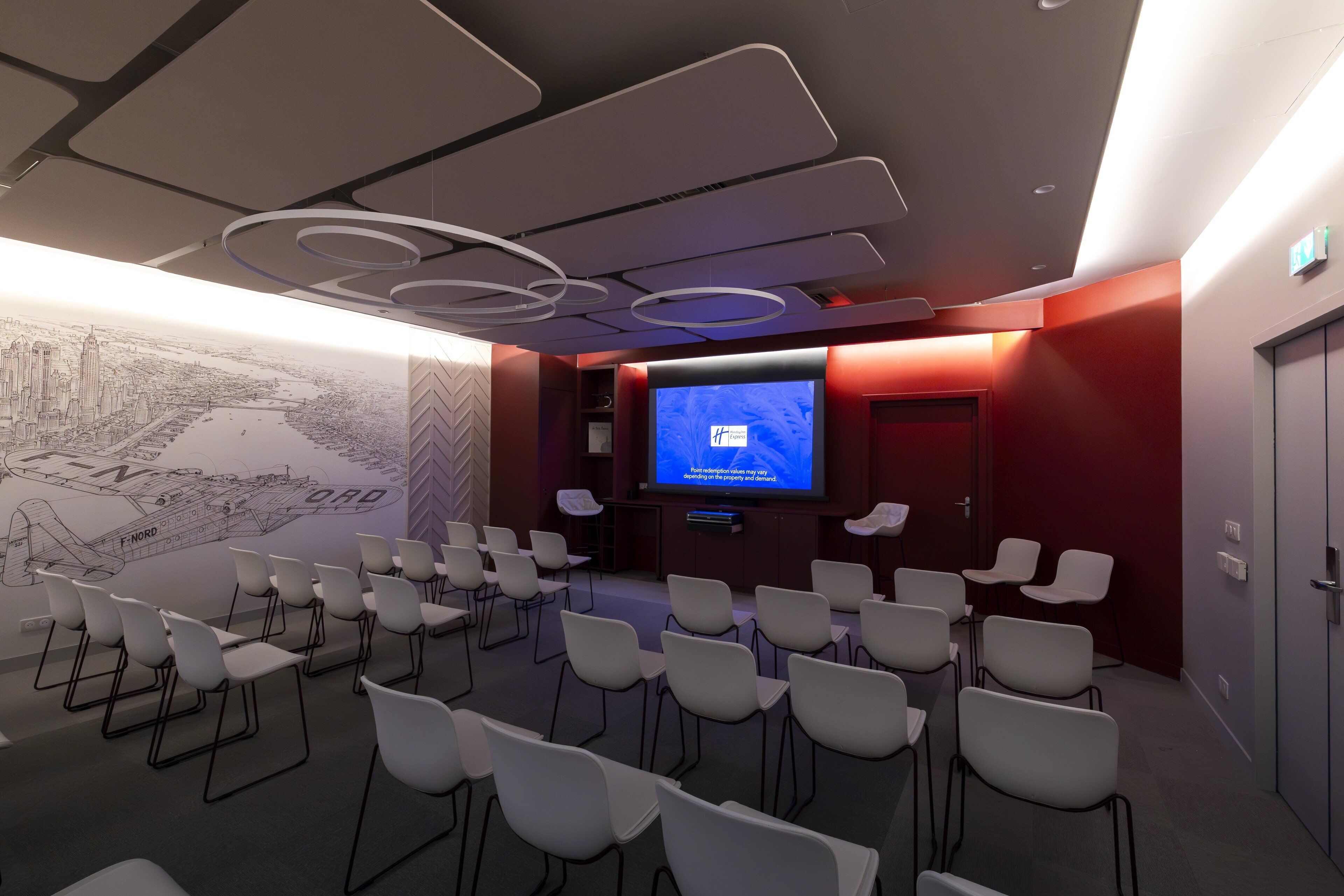 A conference room with rows of white chairs, a large screen, and a cityscape mural on the wall.