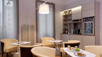 Habitación de hotel con zona de estar que incluye cuatro sillas y dos mesas, un minibar y un ventanal grande con persianas.