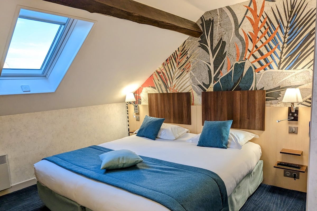 Una habitación de hotel con una cama grande, un tragaluz, un papel tapiz con diseño de hojas, un cabecero de madera y una mesita de noche con lámpara.