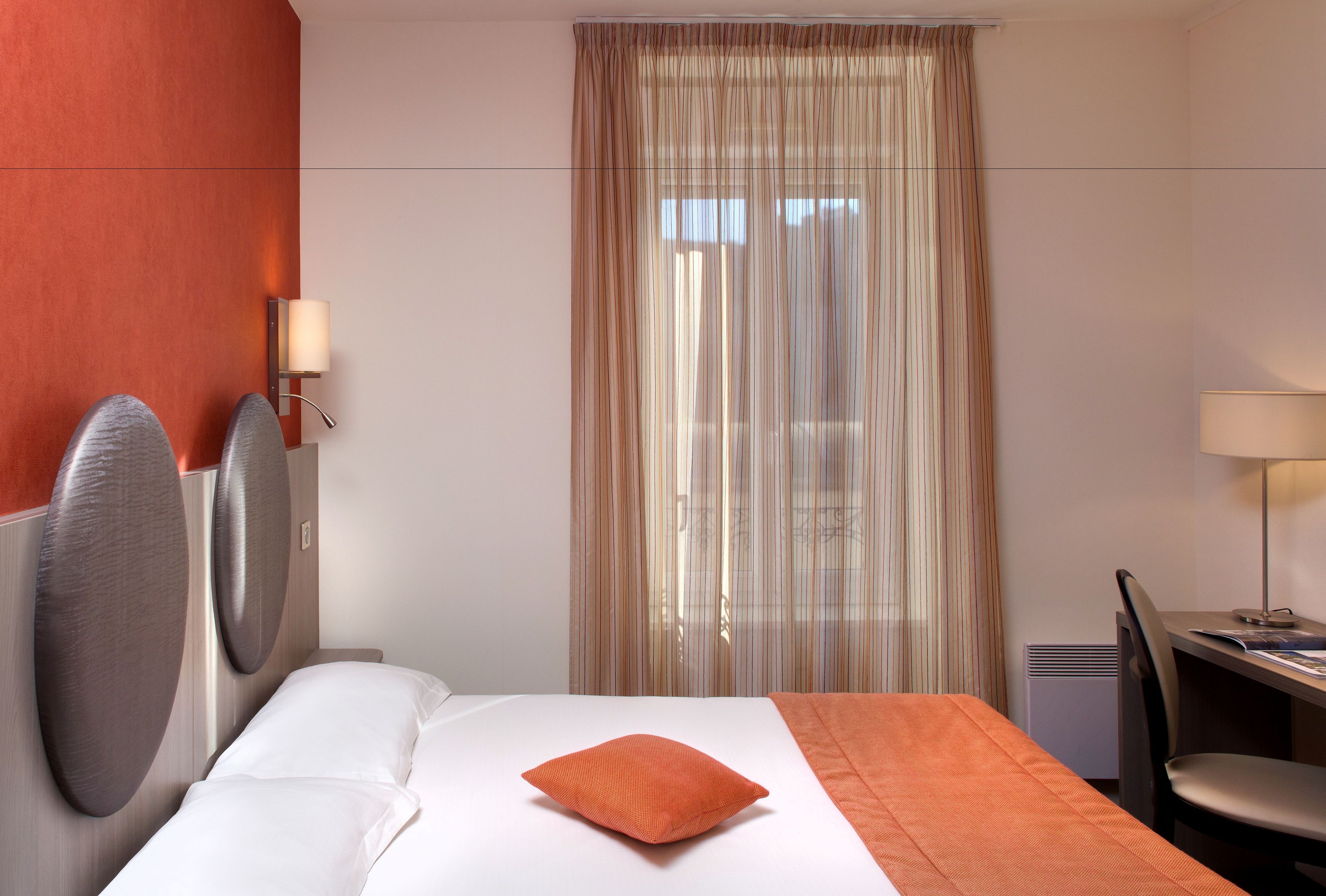 Una habitación de hotel con cama, escritorio, silla, lámpara y ventana con cortinas.
