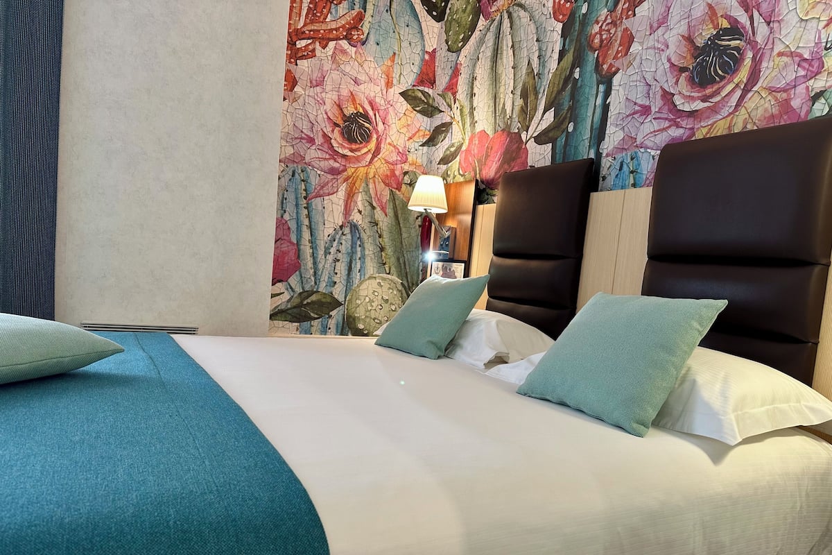Una cama bien hecha con una colcha y almohadas color turquesa, un papel tapiz con motivos florales y un cabecero moderno.