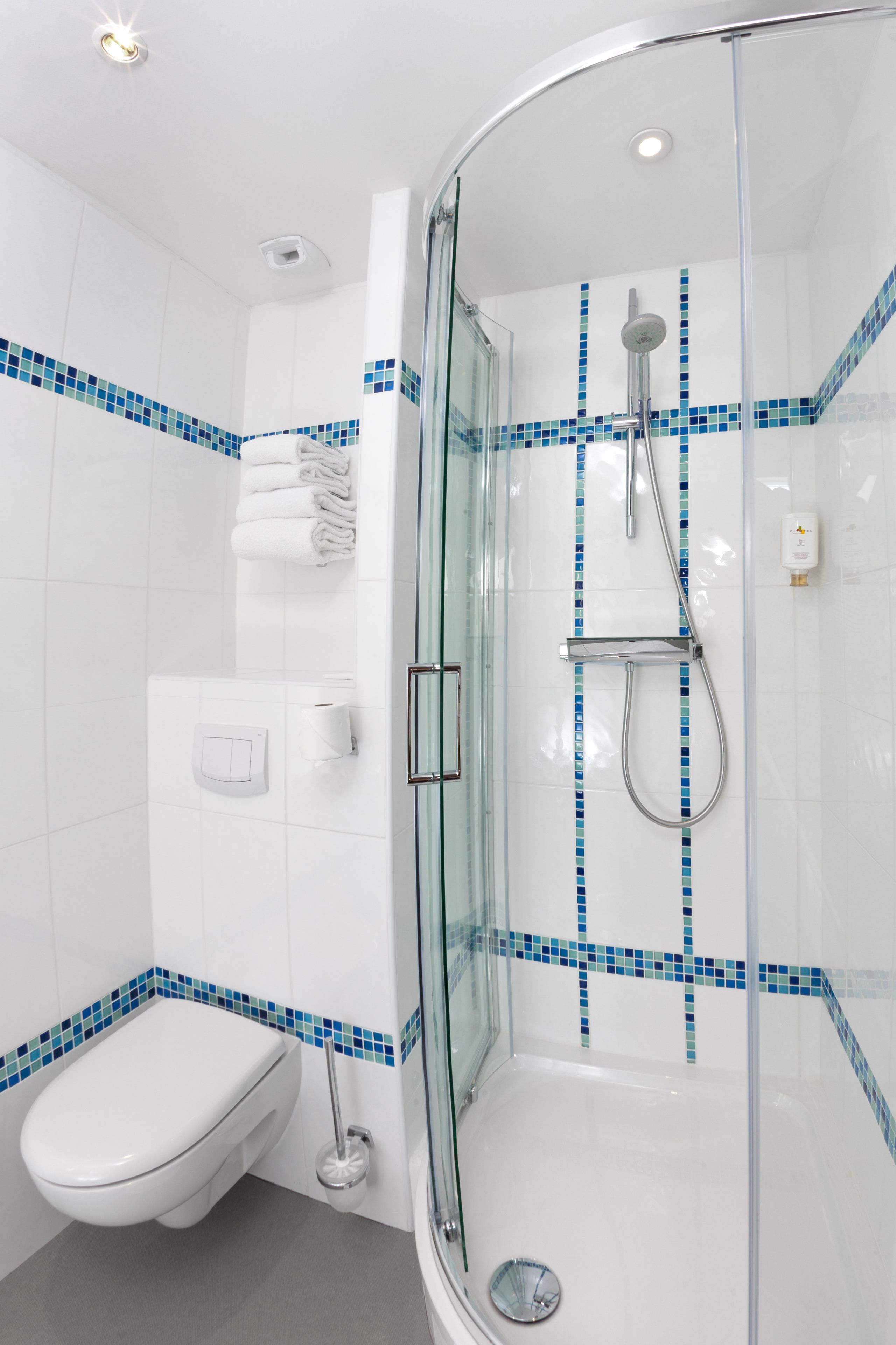 Un baño moderno con ducha de vidrio, azulejos blancos con detalles azules, un inodoro y un perchero para toallas.