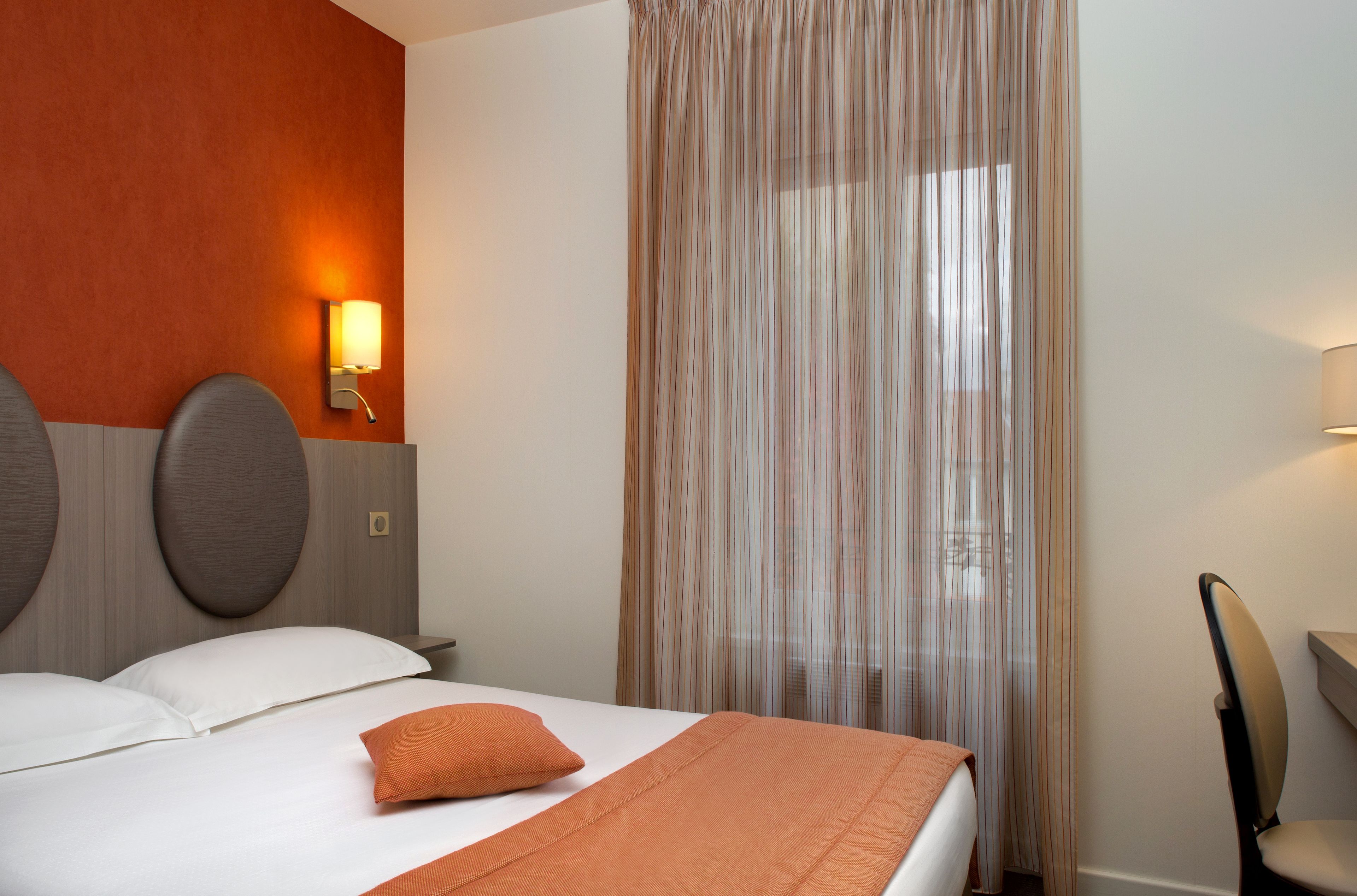 Habitación de hotel con cama, cabecera con lámpara, silla y ventana con cortinas translúcidas.