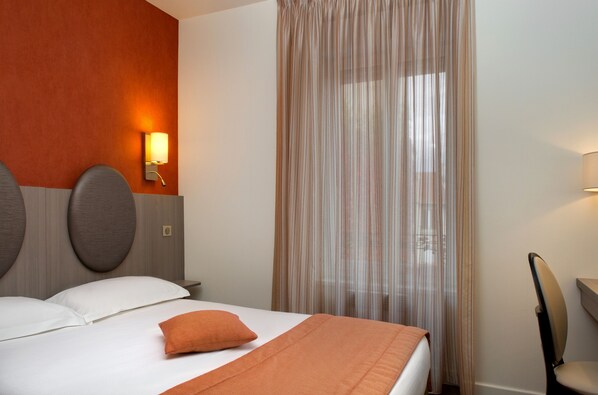 A hotel room with a bed, a headboard with a light fixture, a chair, and a window with sheer curtains.