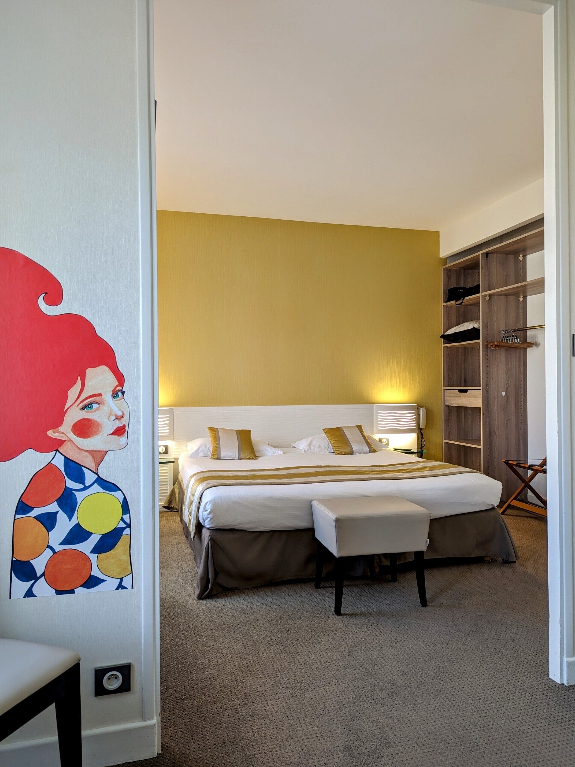 Ein Schlafzimmer mit Bett, Nachttischen, einer Bank, einem Kleiderschrank und einer Wand mit einem farbenfrohen Gemälde.