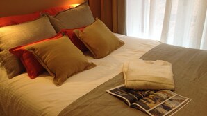 Una cama bien hecha con varias almohadas y una toalla doblada, una revista sobre la cama y una ventana con cortinas.