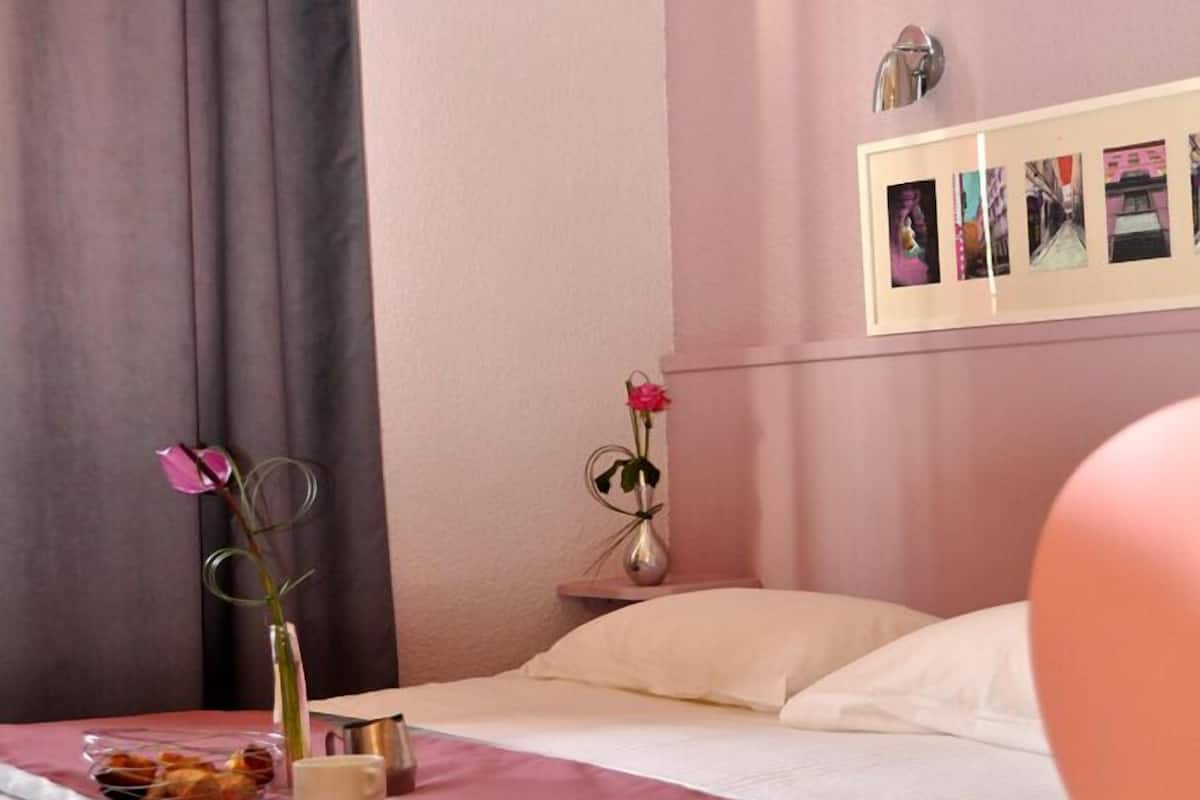 A bedroom with a bed, a nightstand with a vase of flowers, a framed photo collage on the wall, and a window with curtains.