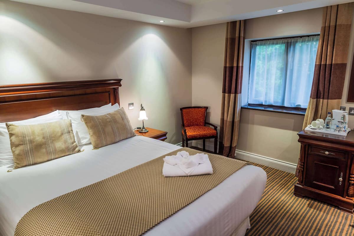 A hotel room with a large bed, a wooden nightstand, a chair, a desk with a lamp, and a window with curtains.