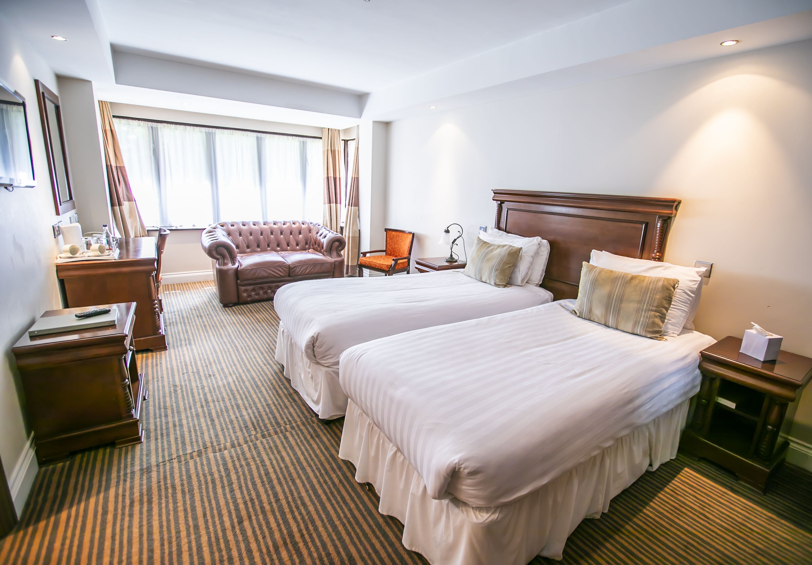 A hotel room with two beds, a sitting area with a chair, a desk, and a large window with curtains.
