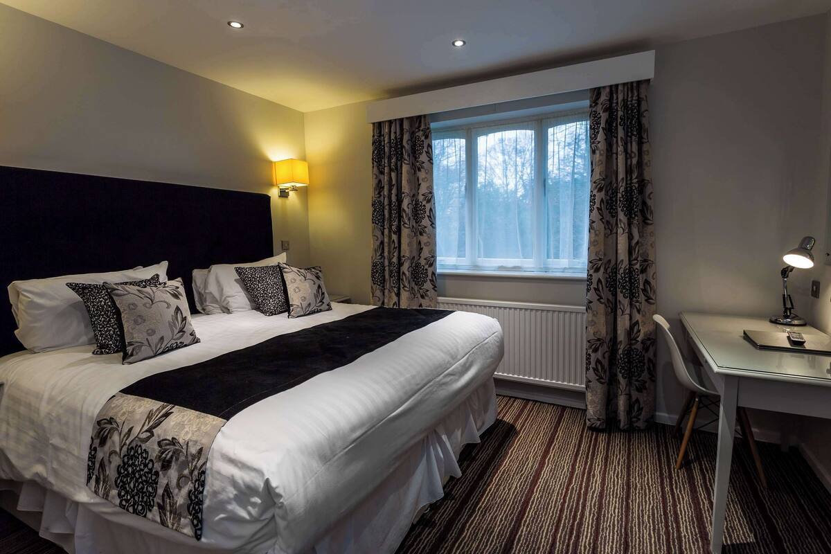 A hotel room with a large bed, a desk, a chair, a window with curtains, and a wall-mounted lamp.
