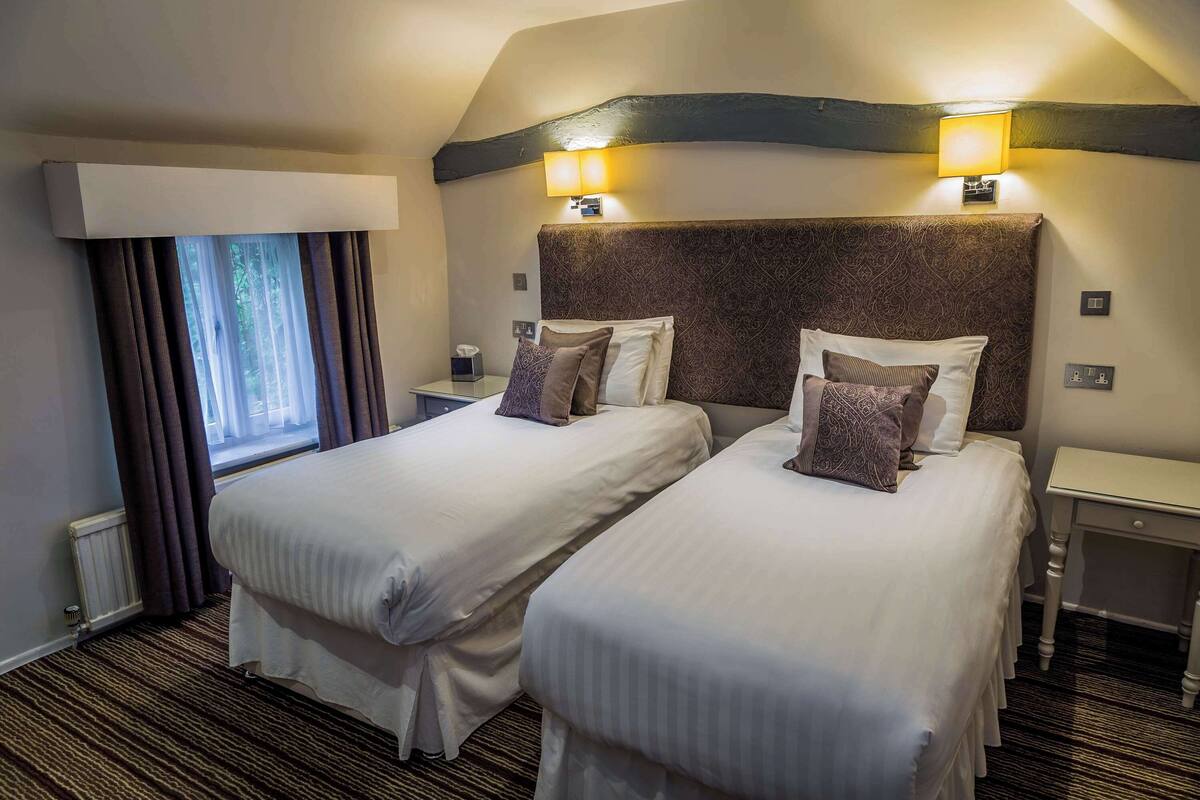 A hotel room with two beds, a window with curtains, a nightstand with a lamp, and a patterned headboard.