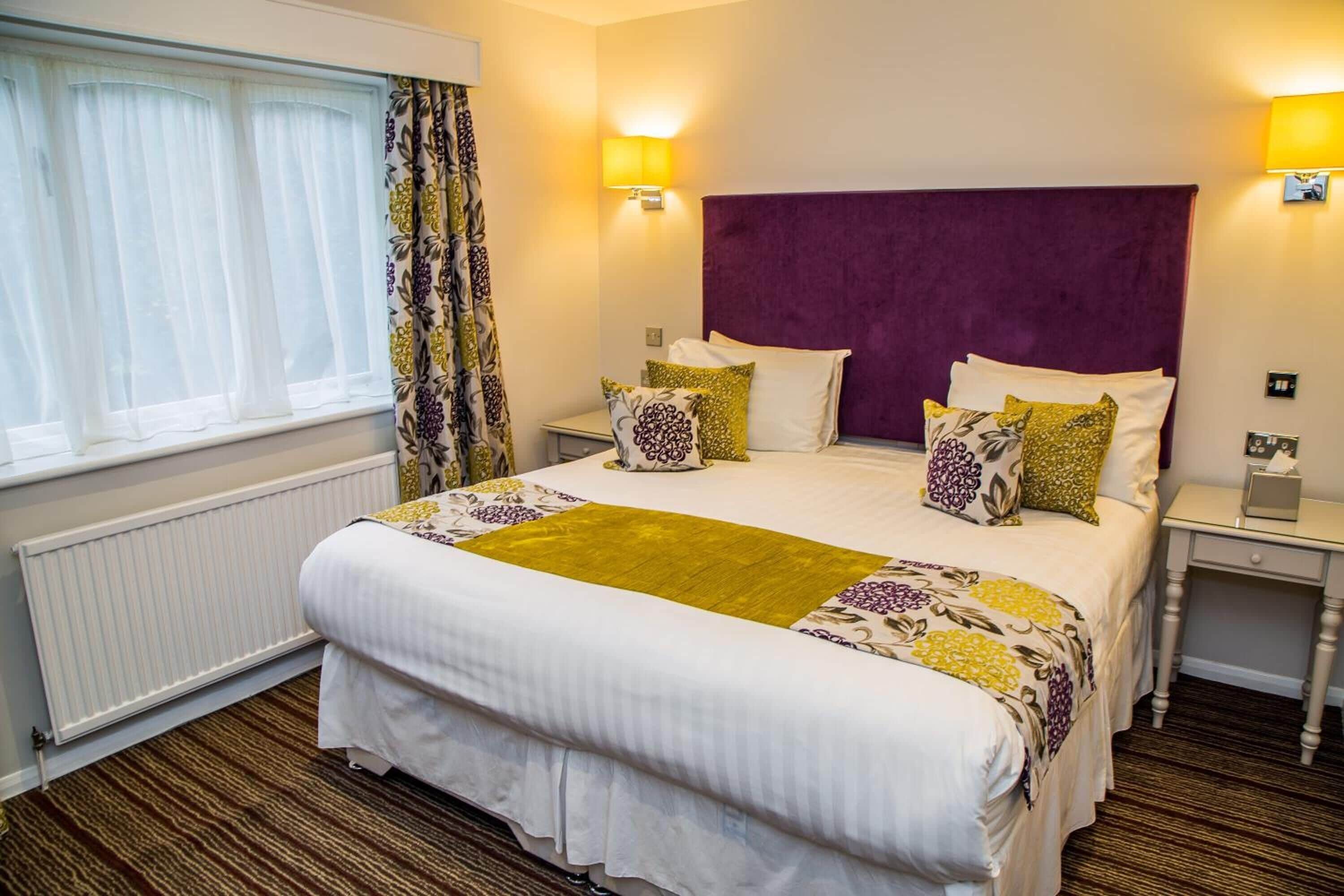 A hotel room with a large bed, a window with sheer curtains, a bedside table with a lamp, and a radiator.