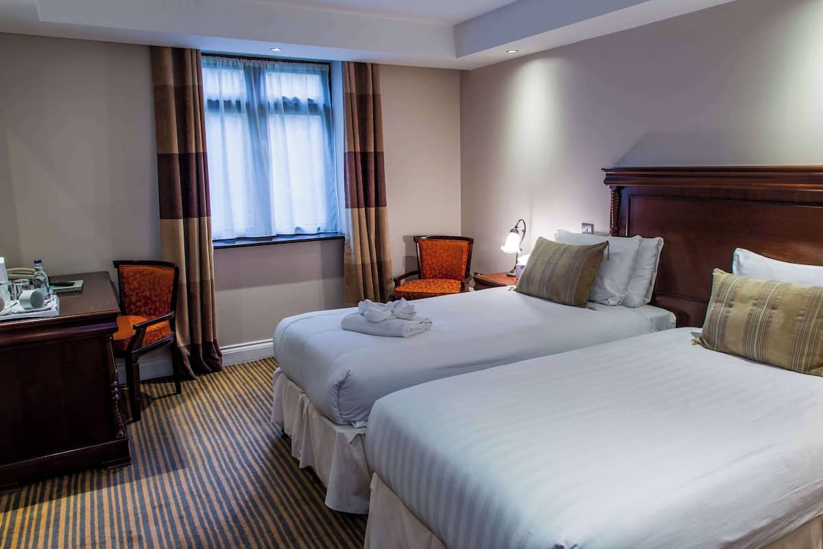 A hotel room with two beds, a desk, a chair, a lamp, and a window with curtains.