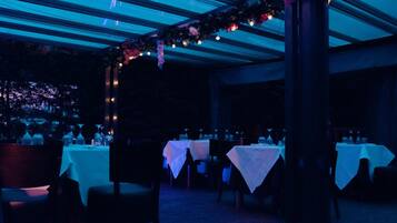 Un área de comedor al aire libre con mesas dispuestas para una comida, bajo un techo cubierto con luces colgantes.