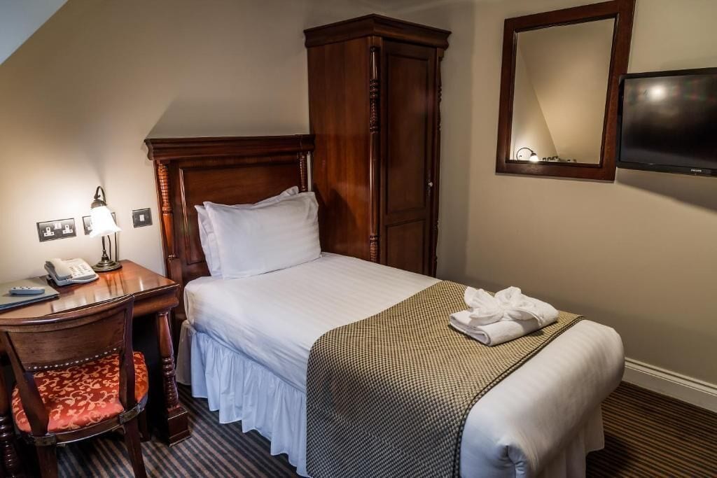 A hotel room with a bed, a wooden wardrobe, a desk with a lamp, a telephone, and a television.