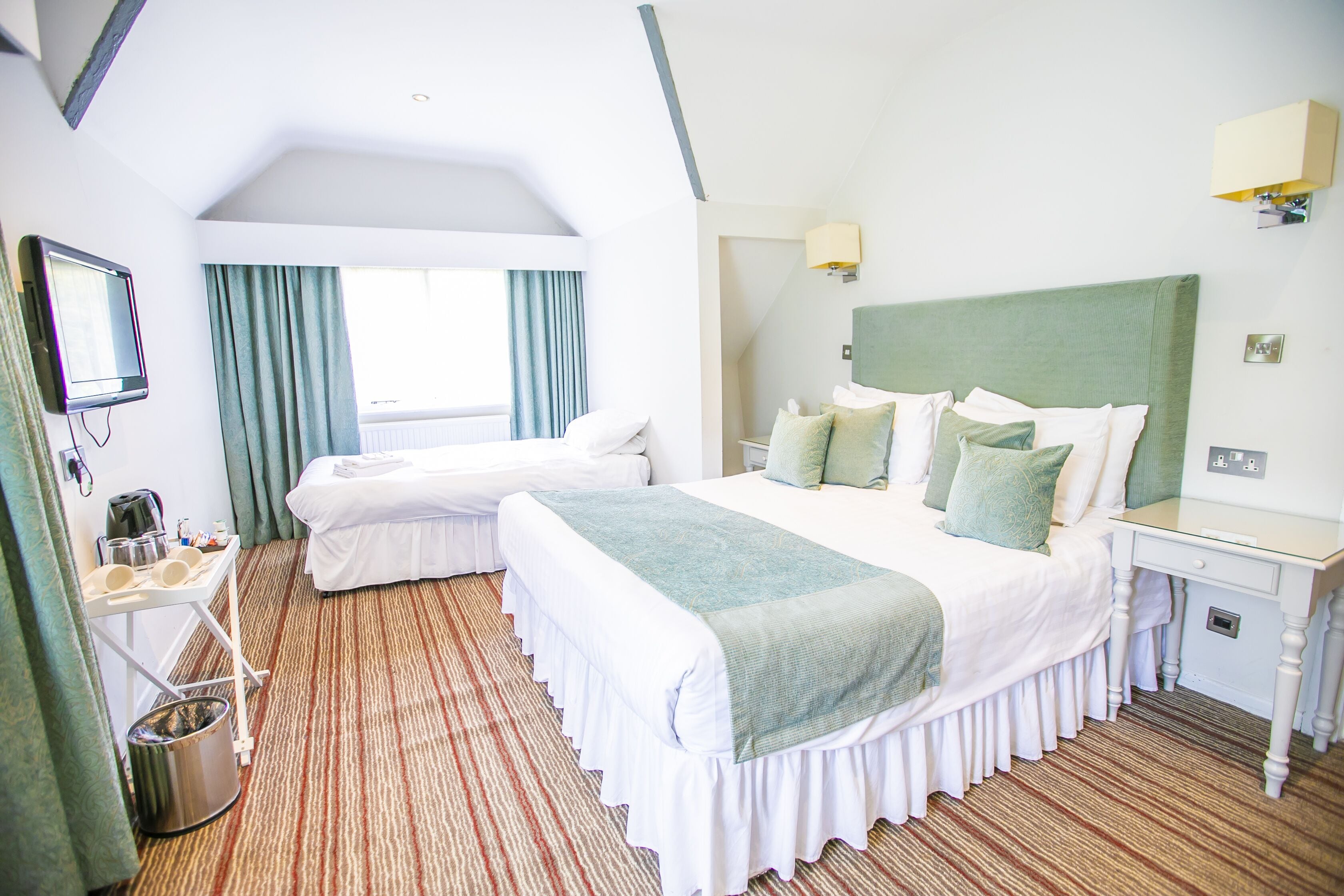 A hotel room with two beds, a small desk, a mirror, a window with curtains, and a radiator.