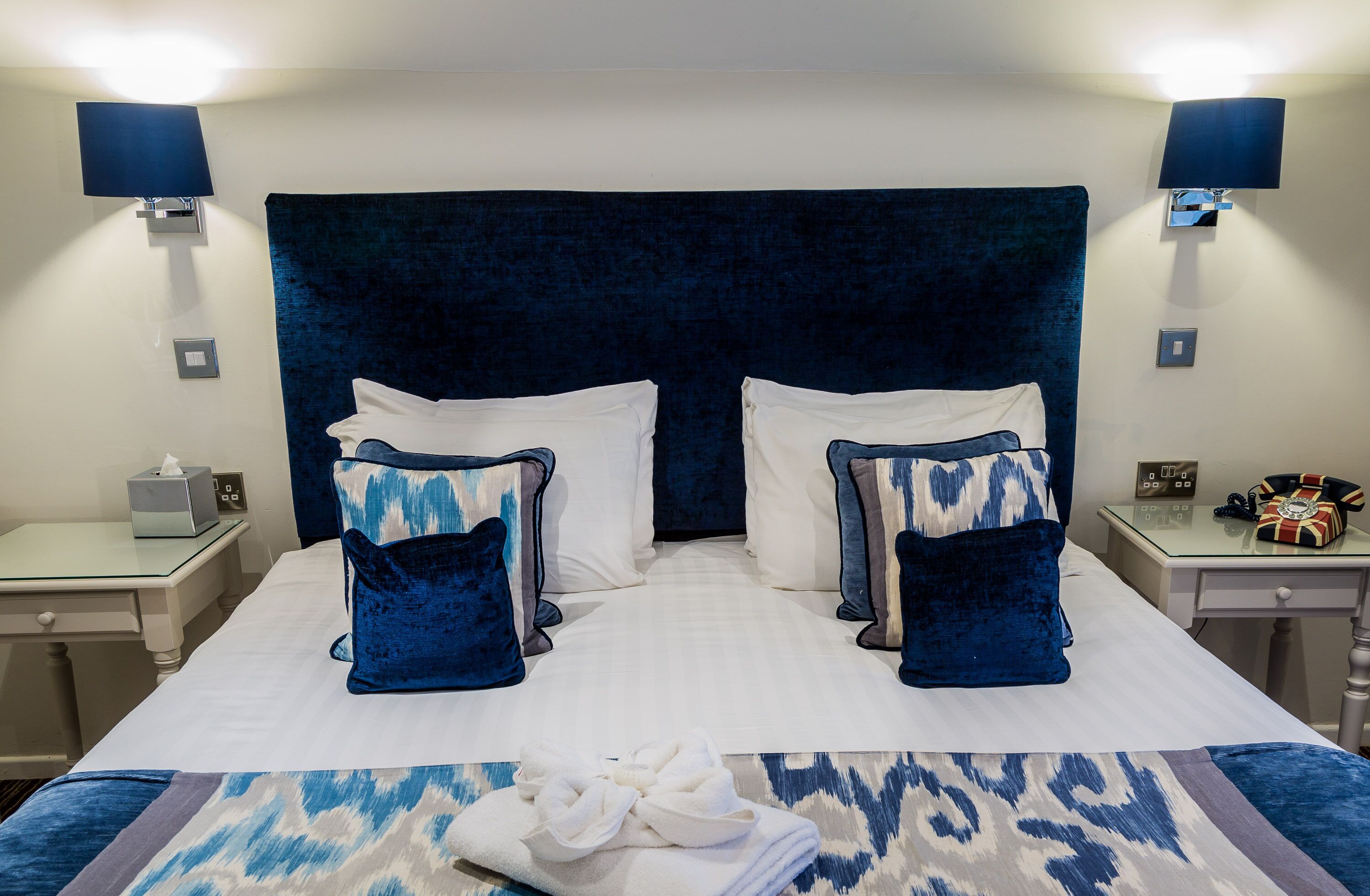 A double bed with a dark headboard, white pillows, and blue patterned pillows. There are bedside tables with lamps and a small clock. A folded towel is on the bed.