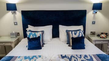 A double bed with a dark headboard, white pillows, and blue patterned pillows. There are bedside tables with lamps and a small clock. A folded towel is on the bed.