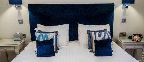 A double bed with a dark headboard, white pillows, and blue patterned pillows. There are bedside tables with lamps and a small clock. A folded towel is on the bed.