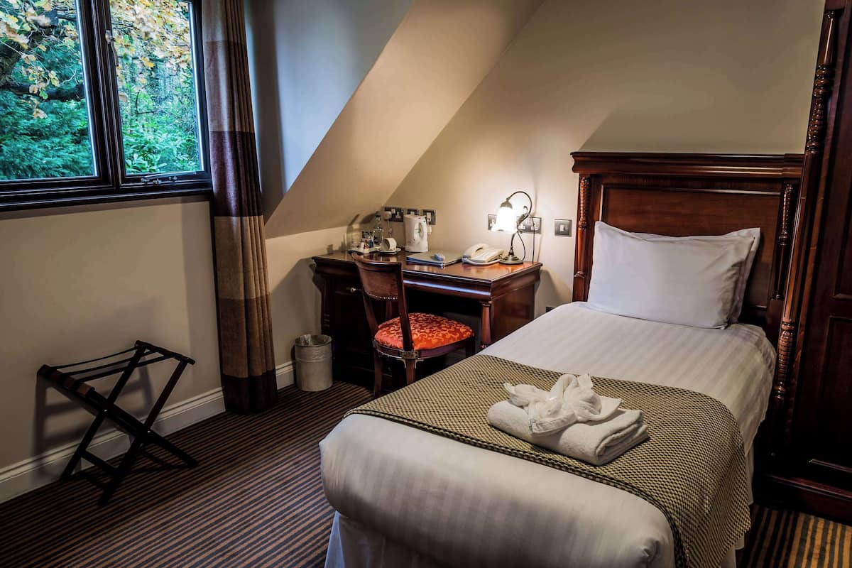 A hotel room with a bed, a desk with a telephone, a chair, and a window with a view of trees.