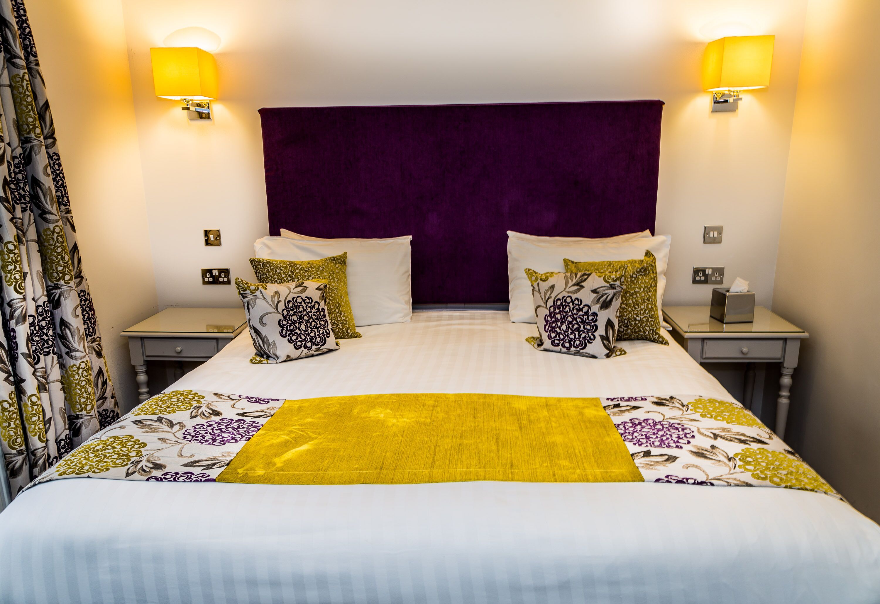 A double bed with a purple headboard, two bedside tables with lamps, and a floral-patterned bedspread.