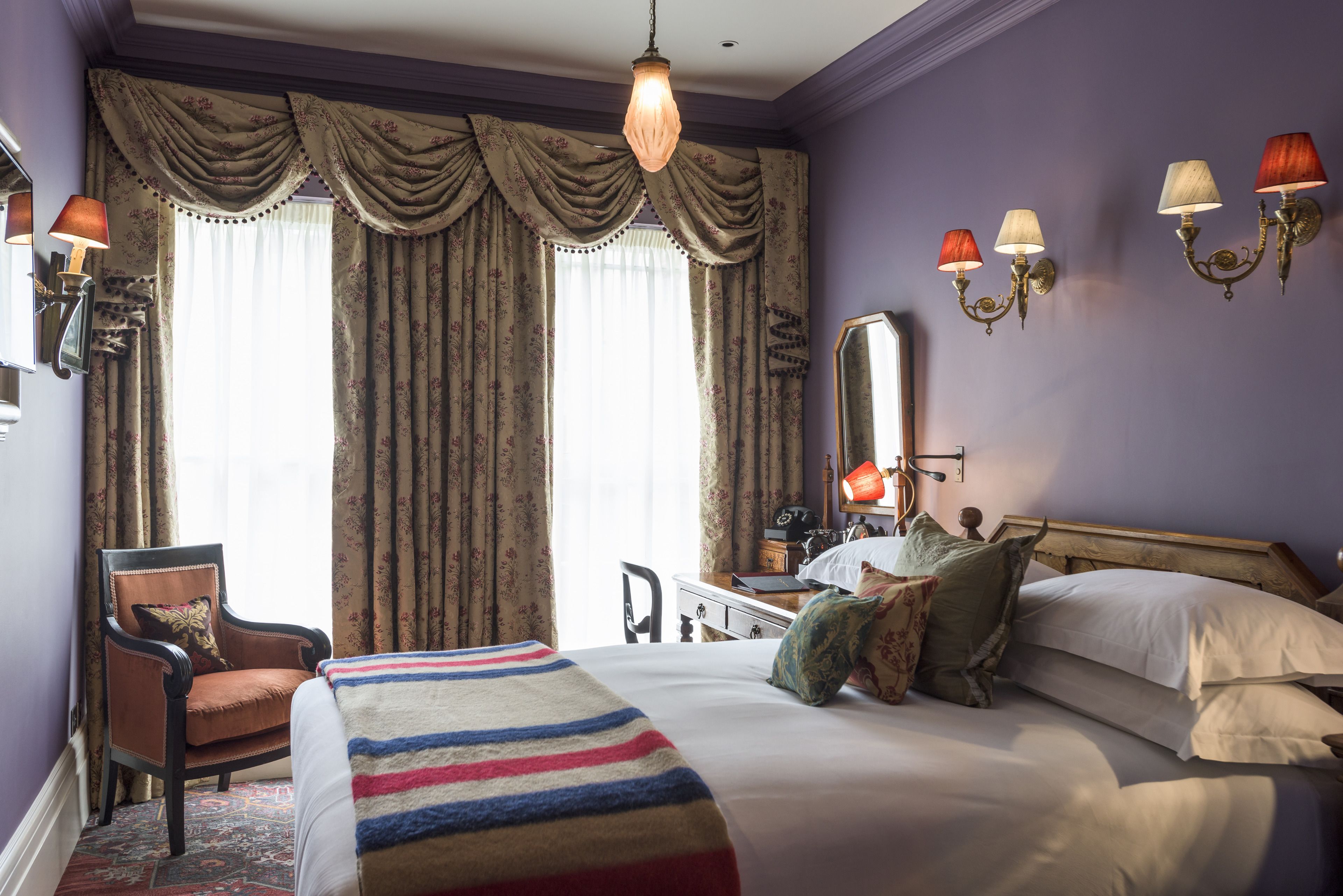A hotel room with a large bed, a chair, a desk with a lamp, and a window with curtains.