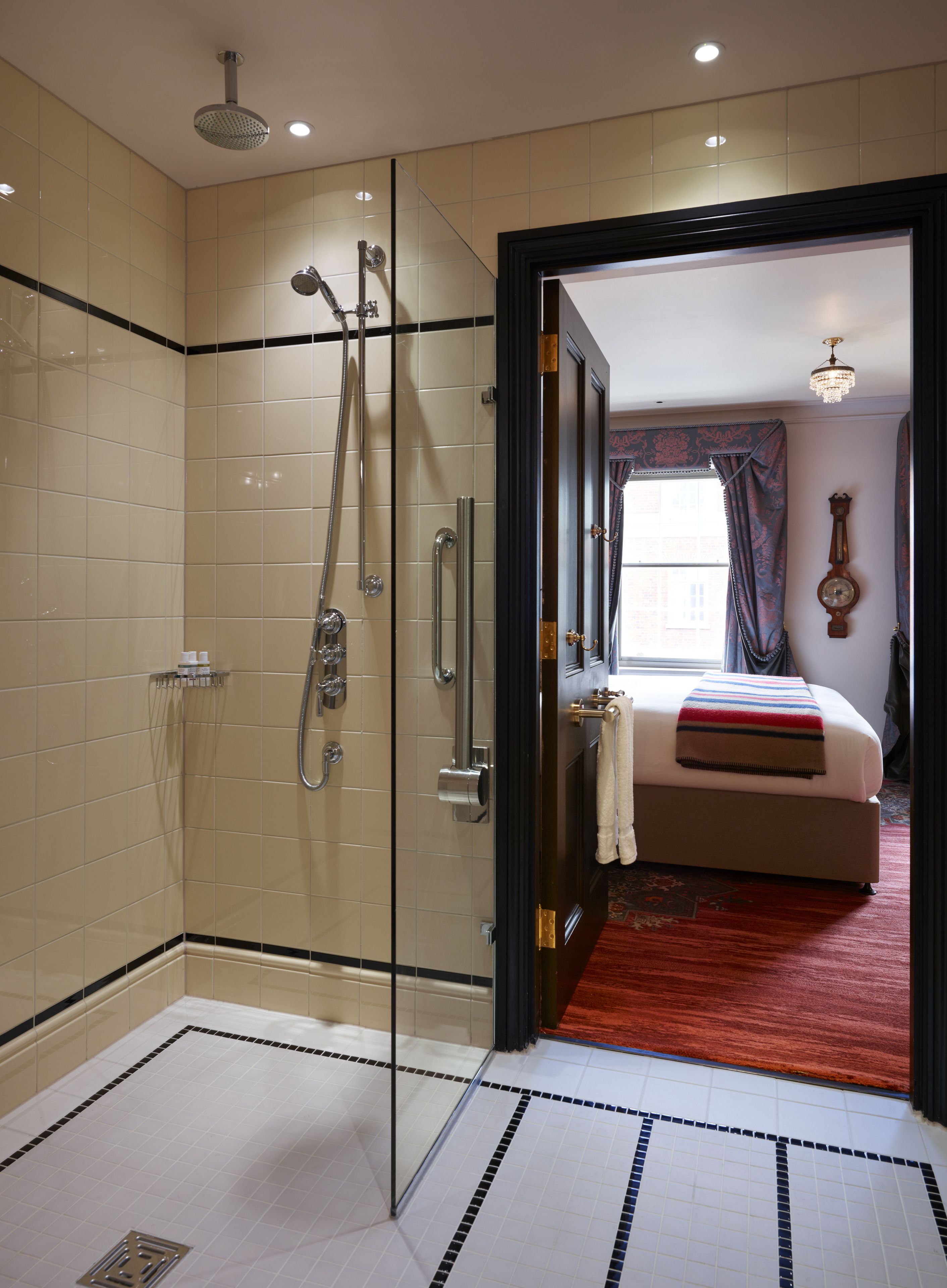 A modern bathroom with a glass-enclosed shower, a visible towel rack, and a visible door leading to a bedroom with a bed and a window.