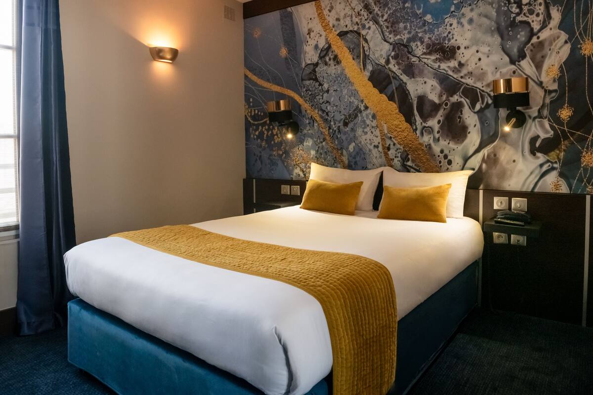 A neatly made bed with a white and yellow bedspread, two bedside tables with lamps, and a wall with a blue abstract design.
