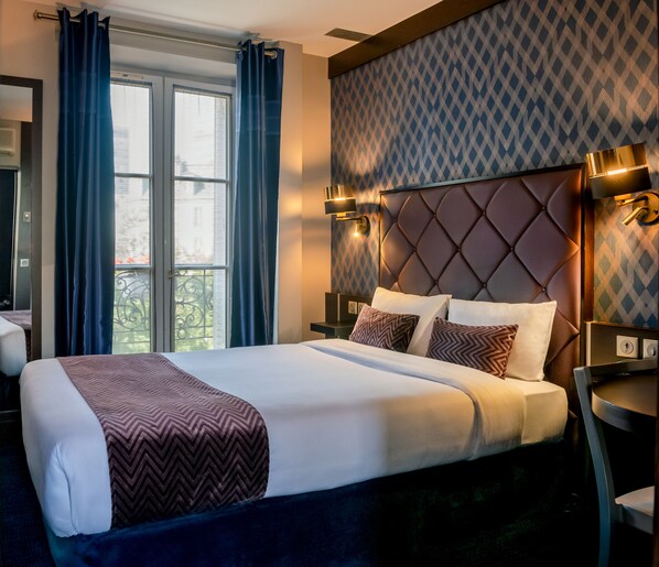 A hotel room with a large bed, a headboard with a diamond pattern, and a window with curtains.