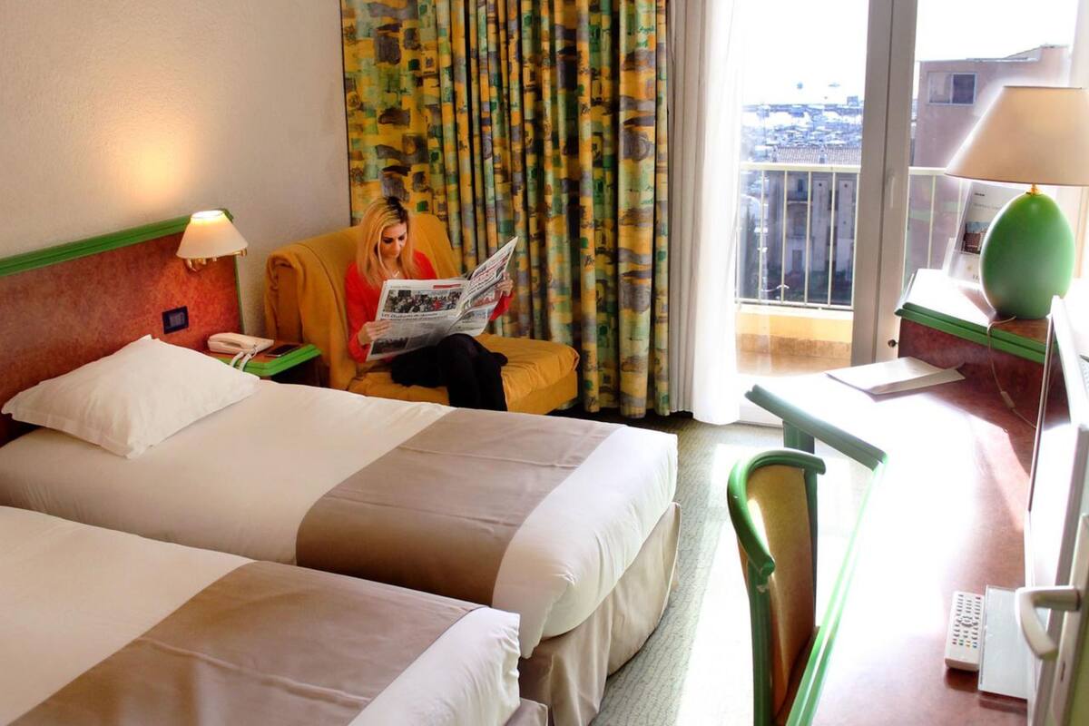 A hotel room with two beds, a chair, a desk with a lamp, and a window with a view of buildings.