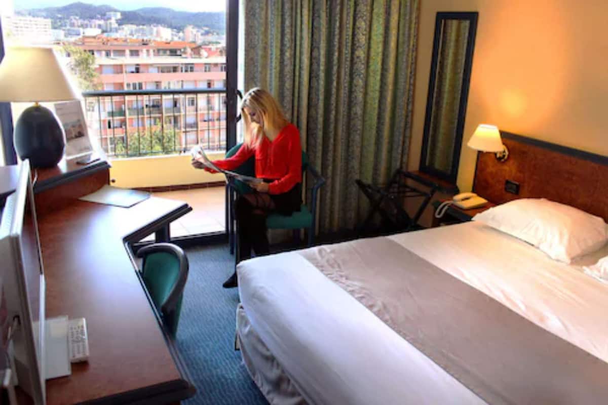 A hotel room with a bed, a desk, a chair, a lamp, and a view of the city.
