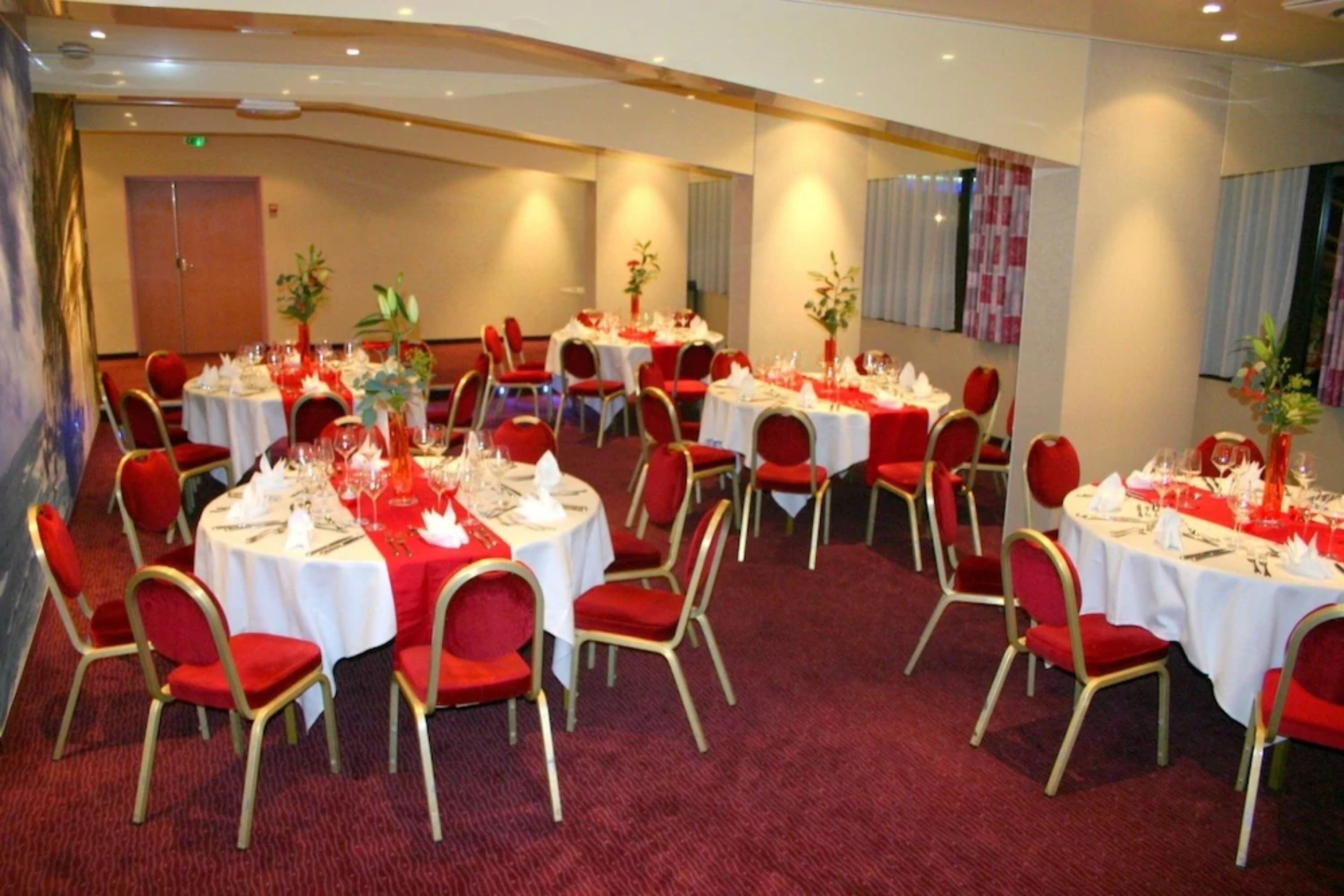 Un salón de banquetes con mesas redondas cubiertas con manteles blancos, sillas rojas y centros de mesa con flores.