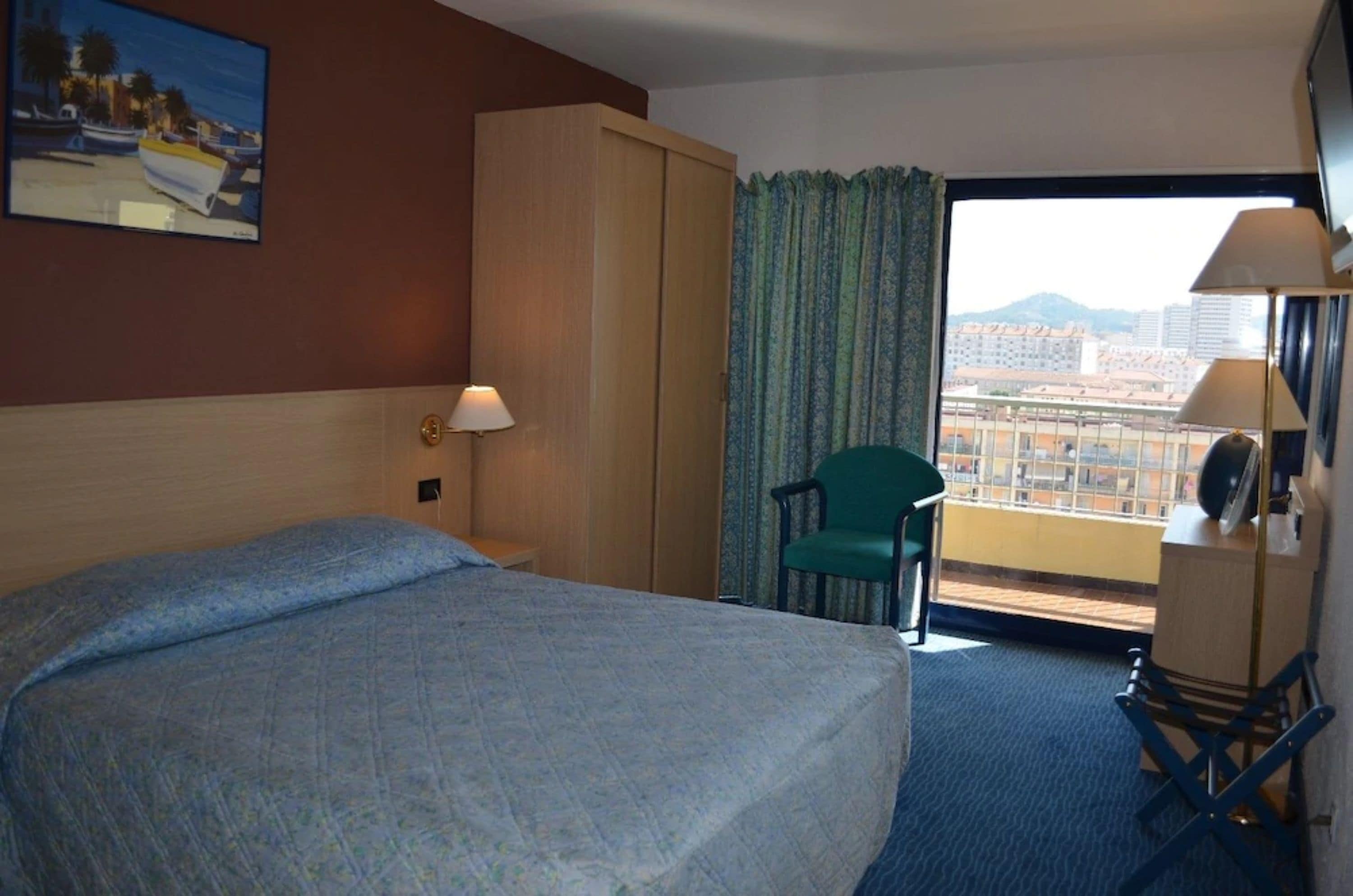 A hotel room with a bed, a chair, a desk, a lamp, and a view of the city through a large window.
