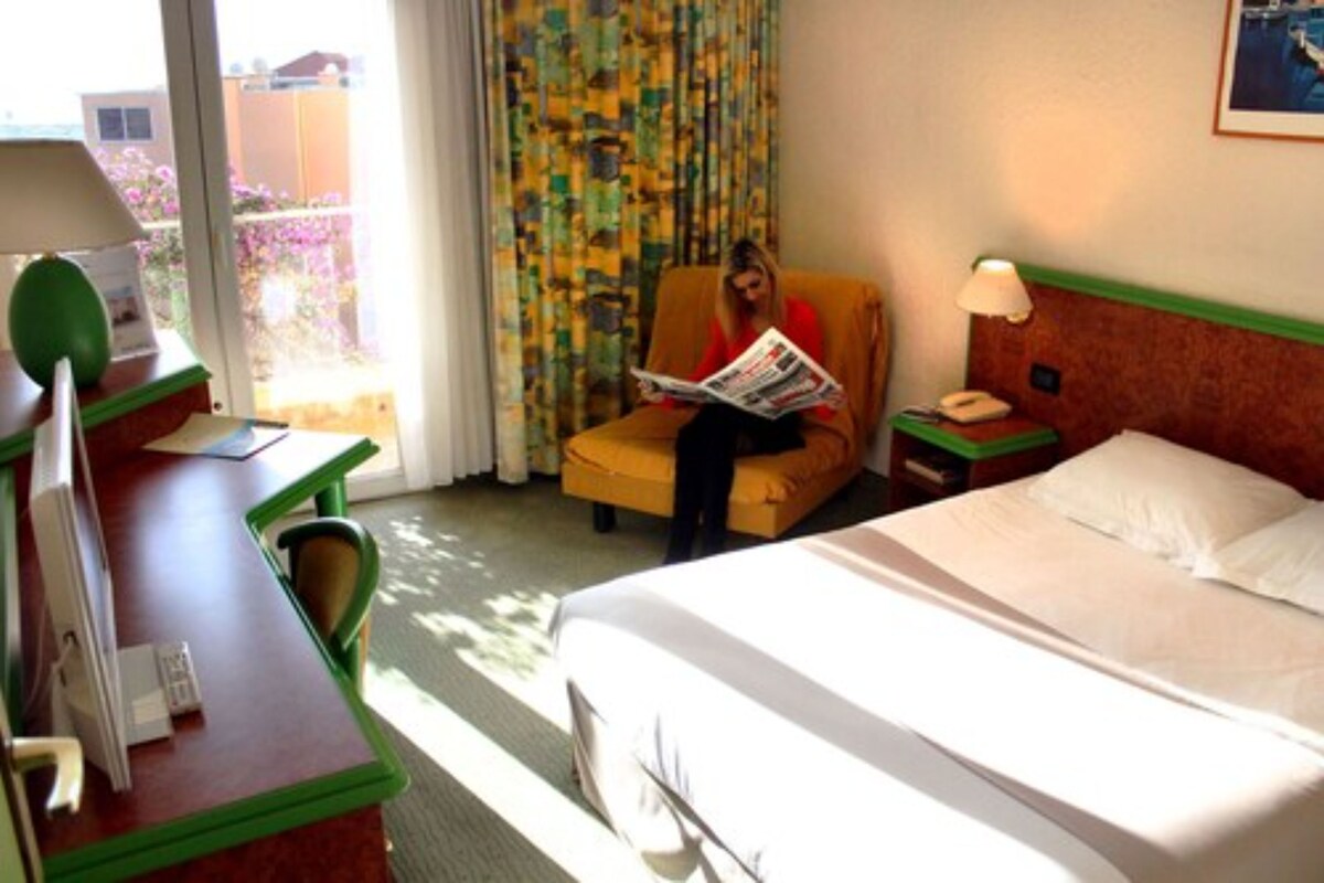 Una habitación de hotel con una cama, un escritorio con silla, un sillón de lectura, una mesita de noche con lámpara y una ventana con cortinas.