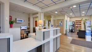 A reception area with a desk, computer, lamp, and a plant. There are bookshelves and a signboard in the background. - Brit Hotel Lille Centre (Lille)