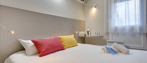 A hotel room with a neatly made bed, a bedside table with a lamp, a wall-mounted air conditioner, and a window with sheer curtains.