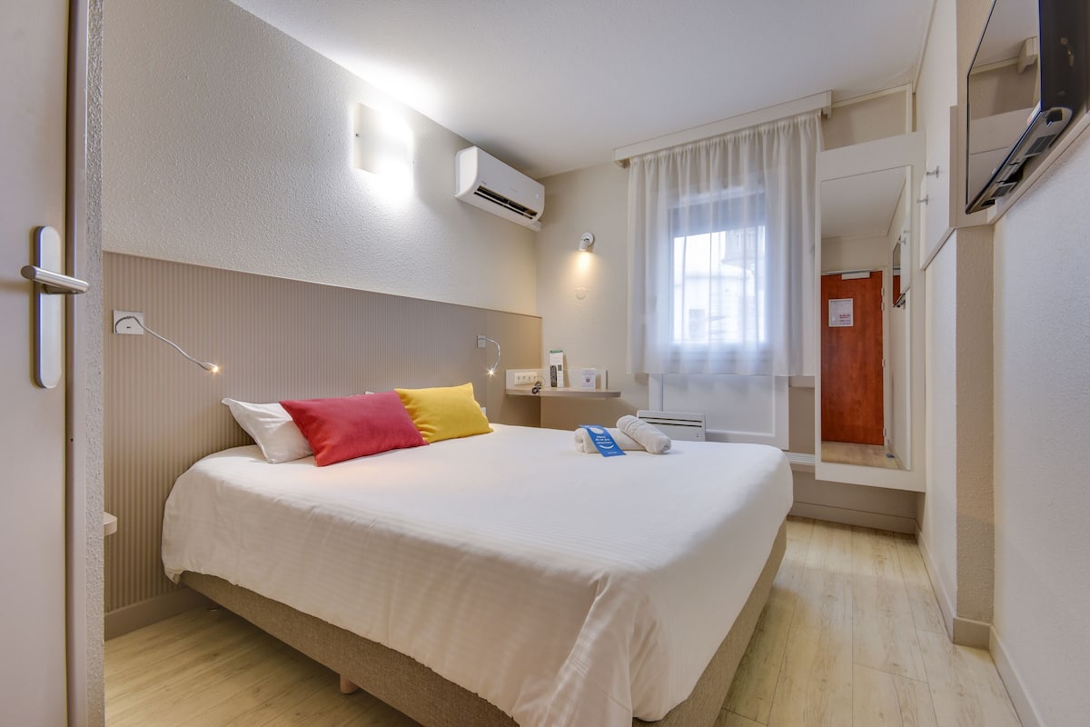 A hotel room with a bed, a bedside table, an air conditioner, a window with curtains, and a door.