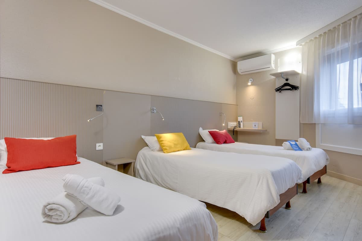 A hotel room with three beds, each with white bedding and different colored pillows. There is a wall-mounted air conditioning unit and a window with curtains.