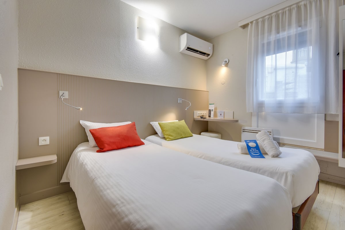 A hotel room with two beds, a window with sheer curtains, an air conditioning unit, and a bedside table with a lamp.