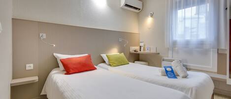 A hotel room with two beds, a window with sheer curtains, an air conditioning unit, and a bedside table with a lamp.