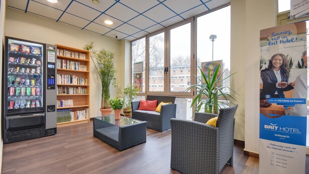 A hotel lobby with a vending machine, bookshelves, wicker furniture, and large windows. - Brit Hotel Lille Centre (Lille)