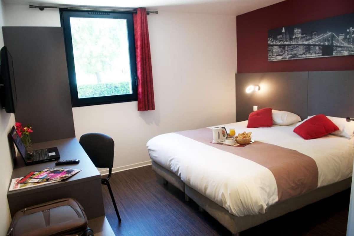 Ein Hotelzimmer mit Bett, Schreibtisch mit Laptop, Stuhl, Fenster mit Vorhang und einem Stadtbild an der Wand.