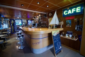 A cafe interior with a wooden counter, bar stools, a menu board, and a neon sign. - Le Voltaire (Rennes)