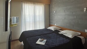 A hotel room with two beds, a flat-screen TV, a wooden headboard, a window with curtains, and a ceiling light. - Le Voltaire (Rennes)