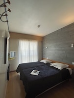 Une chambre d’hôtel avec deux lits, un téléviseur à écran plat, une tête de lit en bois, une fenêtre avec des rideaux et un luminaire au plafond.