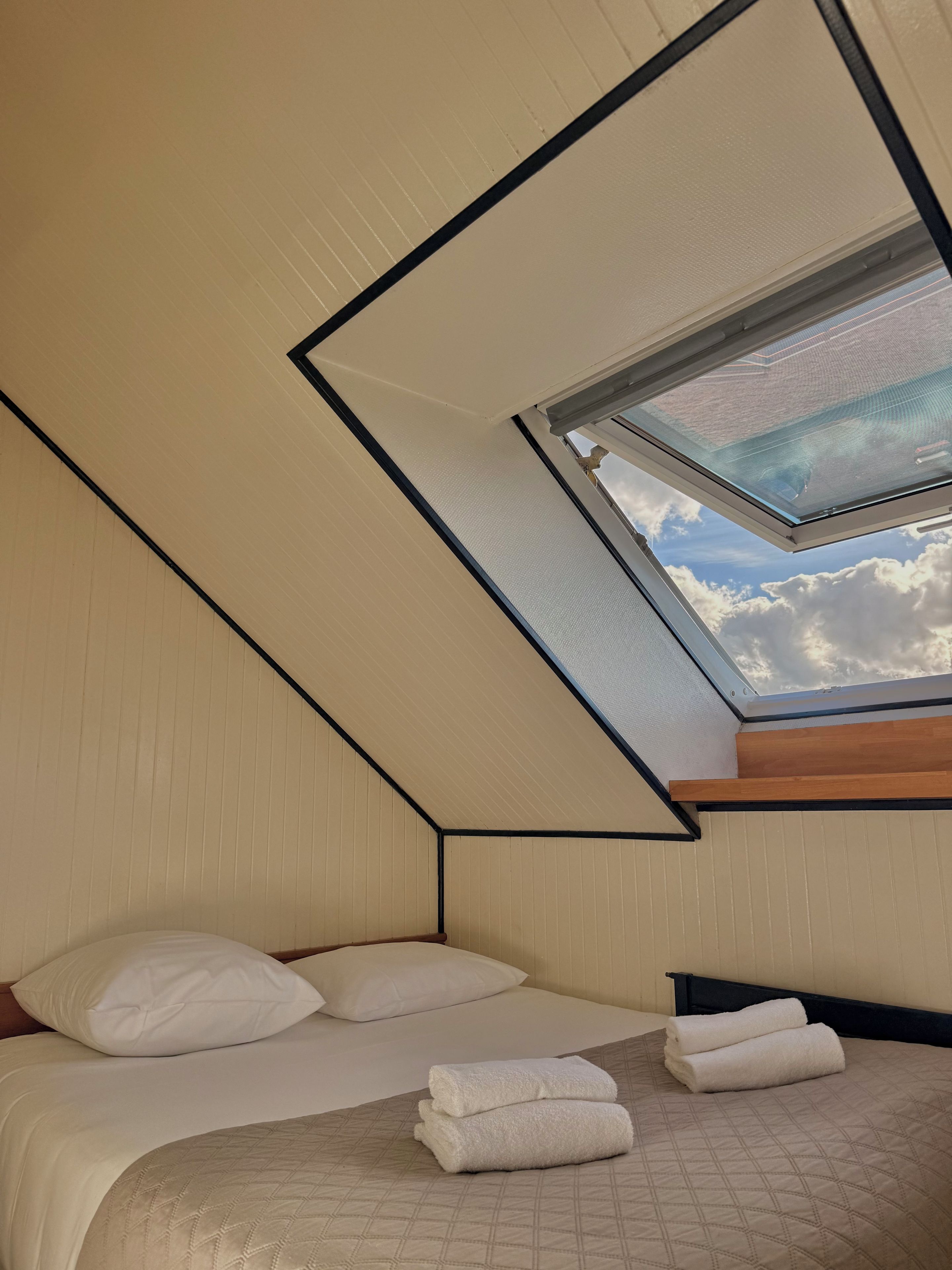A bedroom with a sloped ceiling, a skylight, a bed with white bedding, and a wooden headboard.