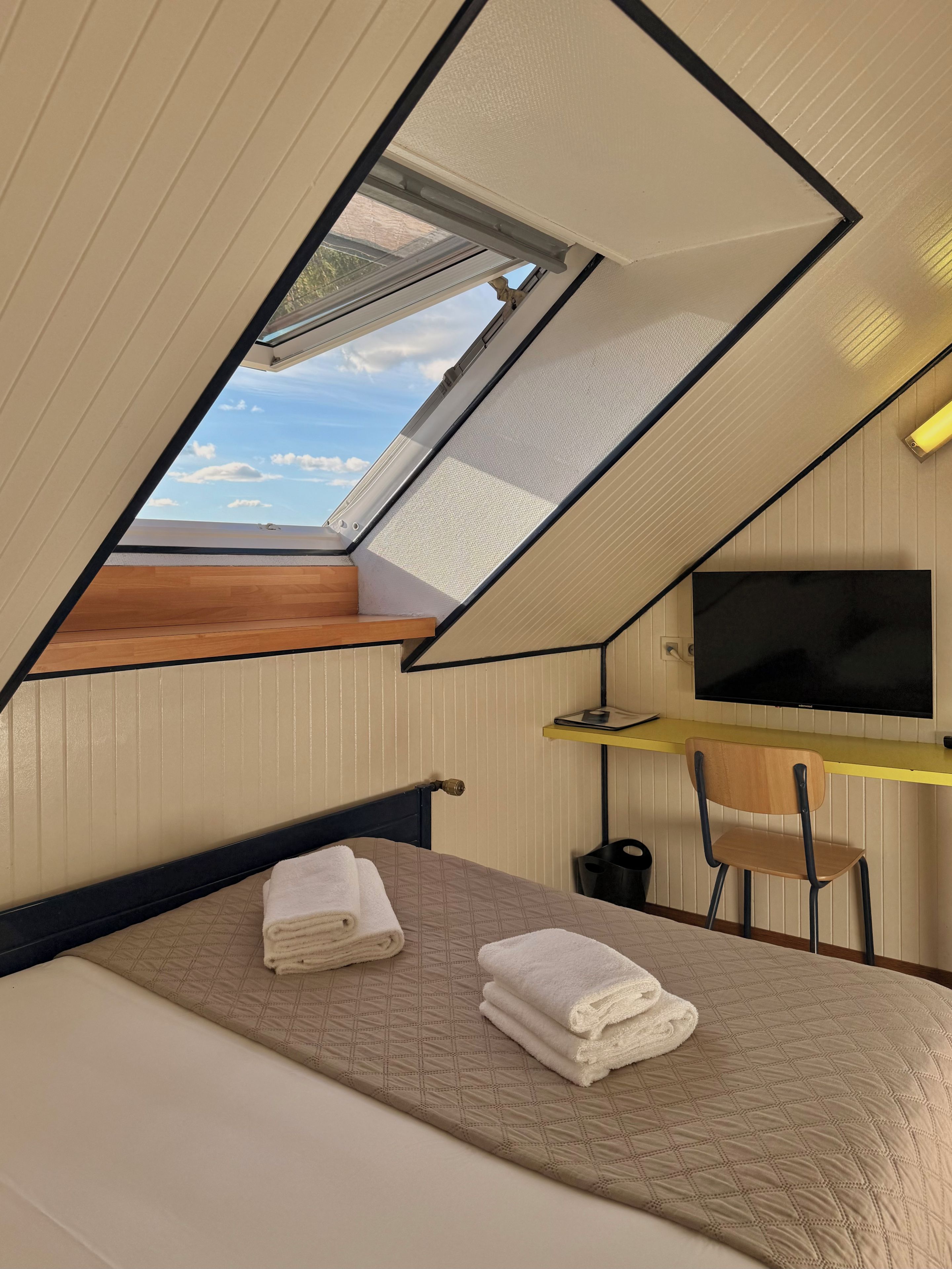 Una habitación con cama, un escritorio con silla, televisión y tragaluz.